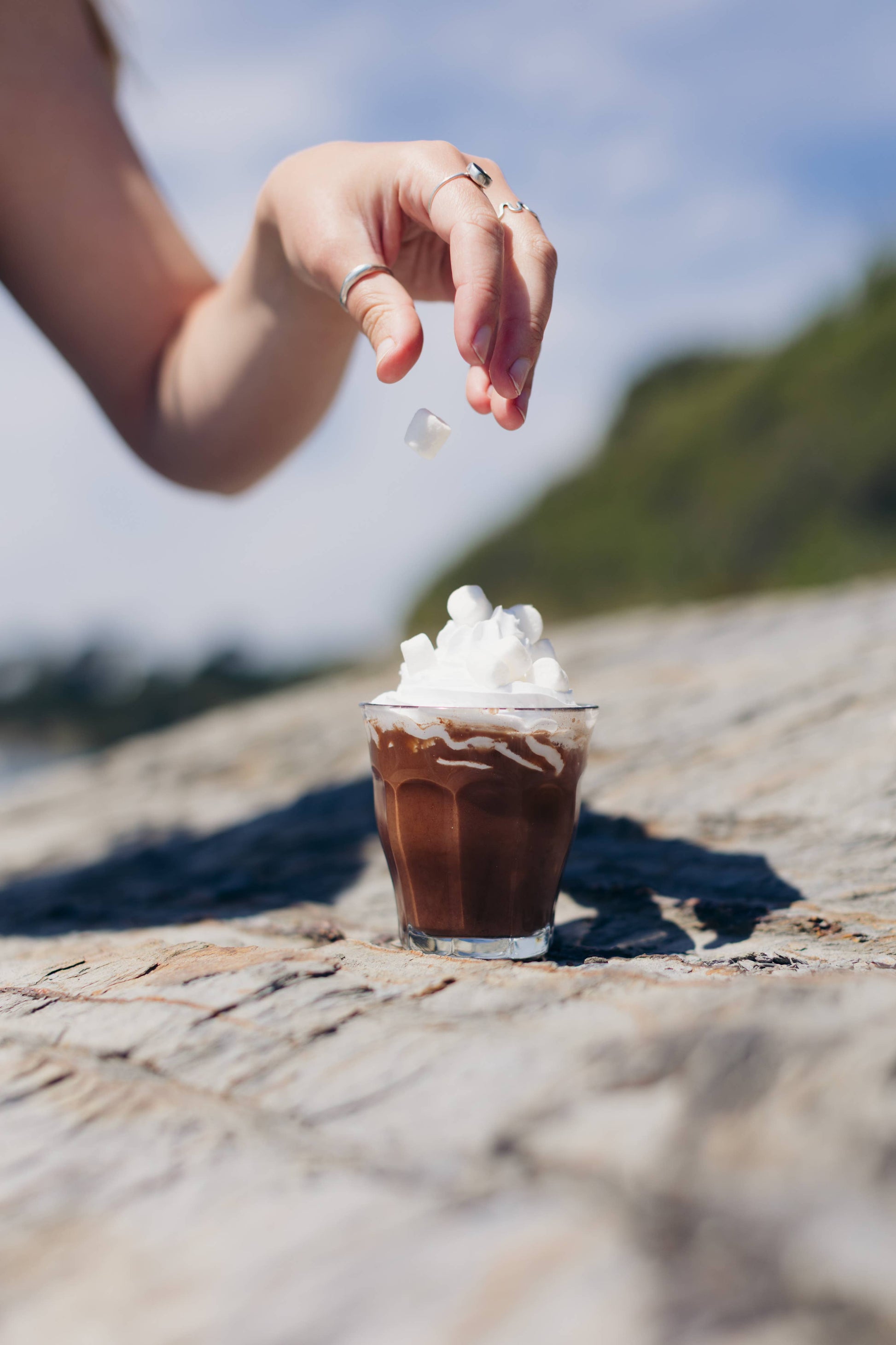 Hand sprinkling a white substance onto a small glass of iced coffee with whipped cream on a stone surface.