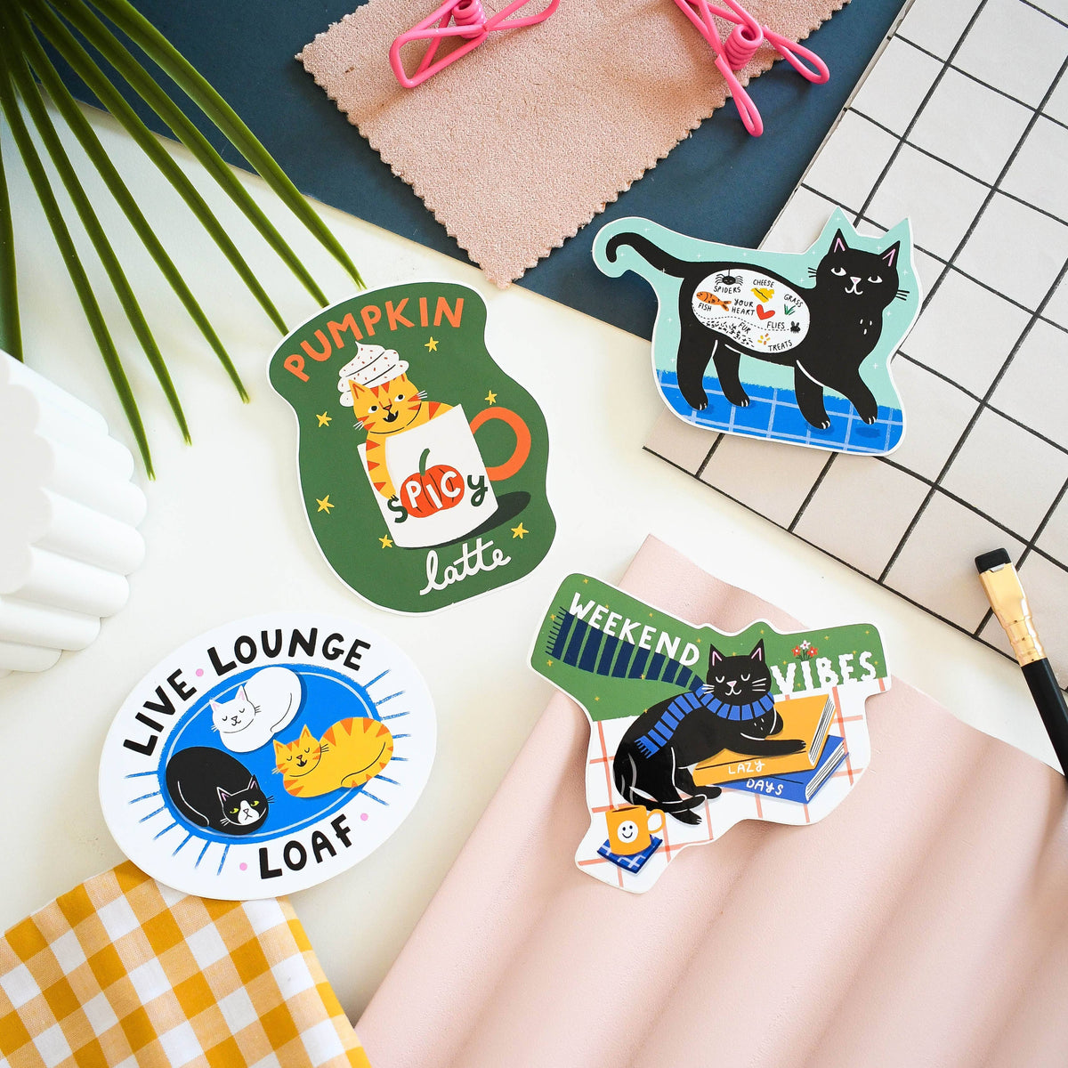 Four die-cut sticker designs featuring cartoon cats in different scenes, arranged on a styled flat-lay surface with pink paper, a green plant leaf, a checkered notebook, and a pen.