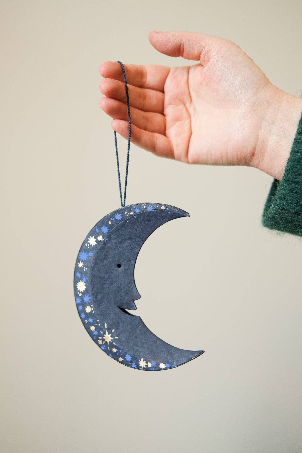 Hand holding a blue crescent moon ornament with star and moon designs against a beige background