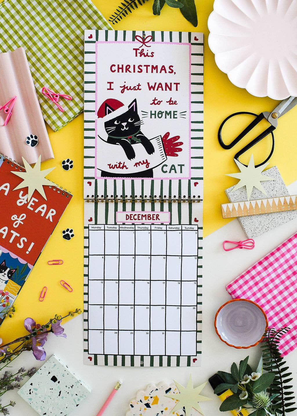 A spiral-bound monthly calendar page with a Christmas-themed cover reading 'This Christmas, I just want to be home with my cat' and a December grid below, styled in a flat lay with paper goods, scissors, star cutouts, and decorative props on a yellow surface.