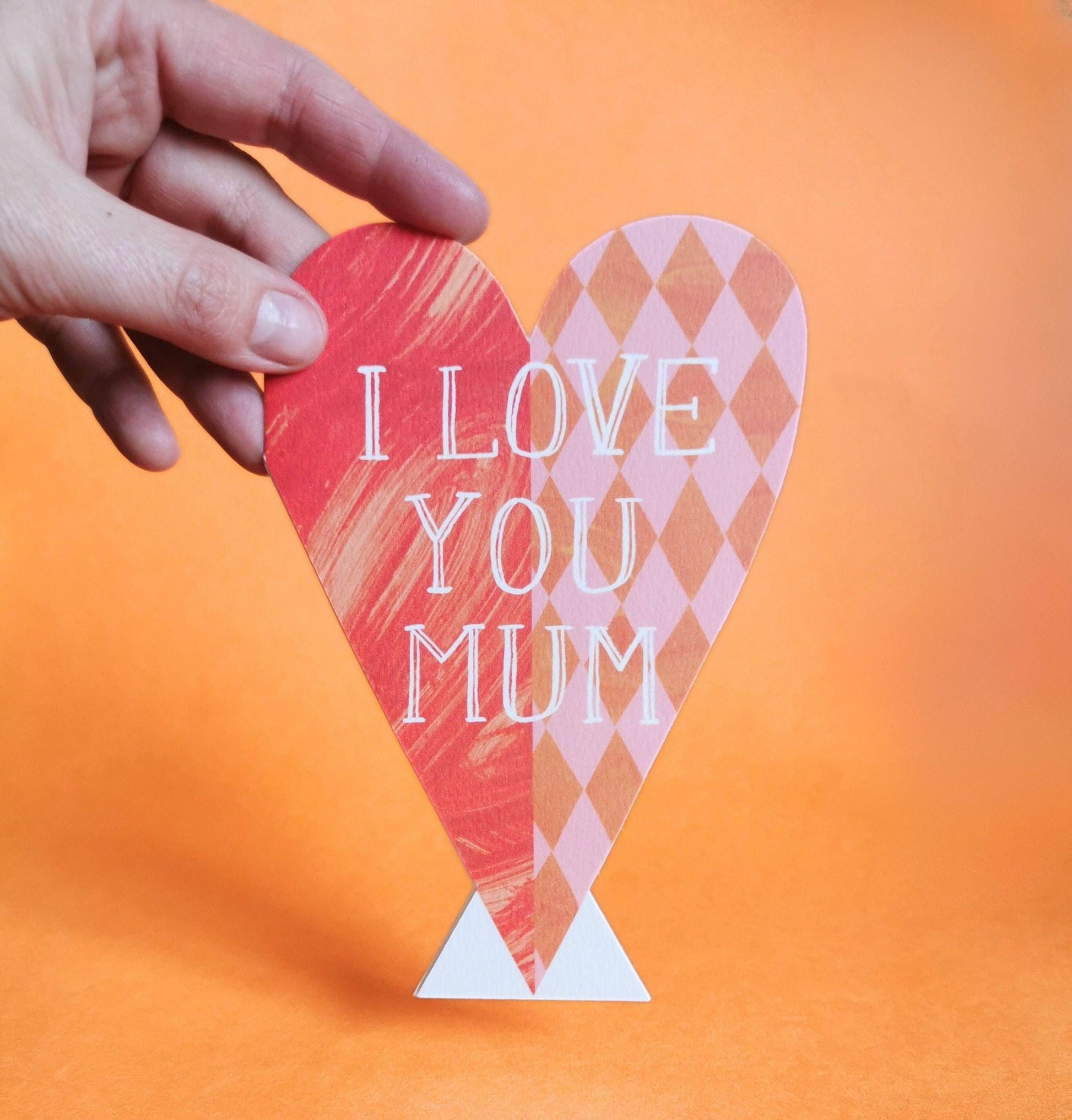 Heart-shaped card with 'I Love You Mum' text held by a hand against an orange background