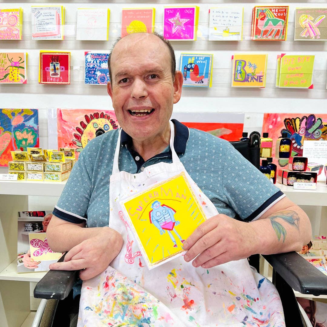 Man in apron holding a colorful card in front of a display of greeting cards.