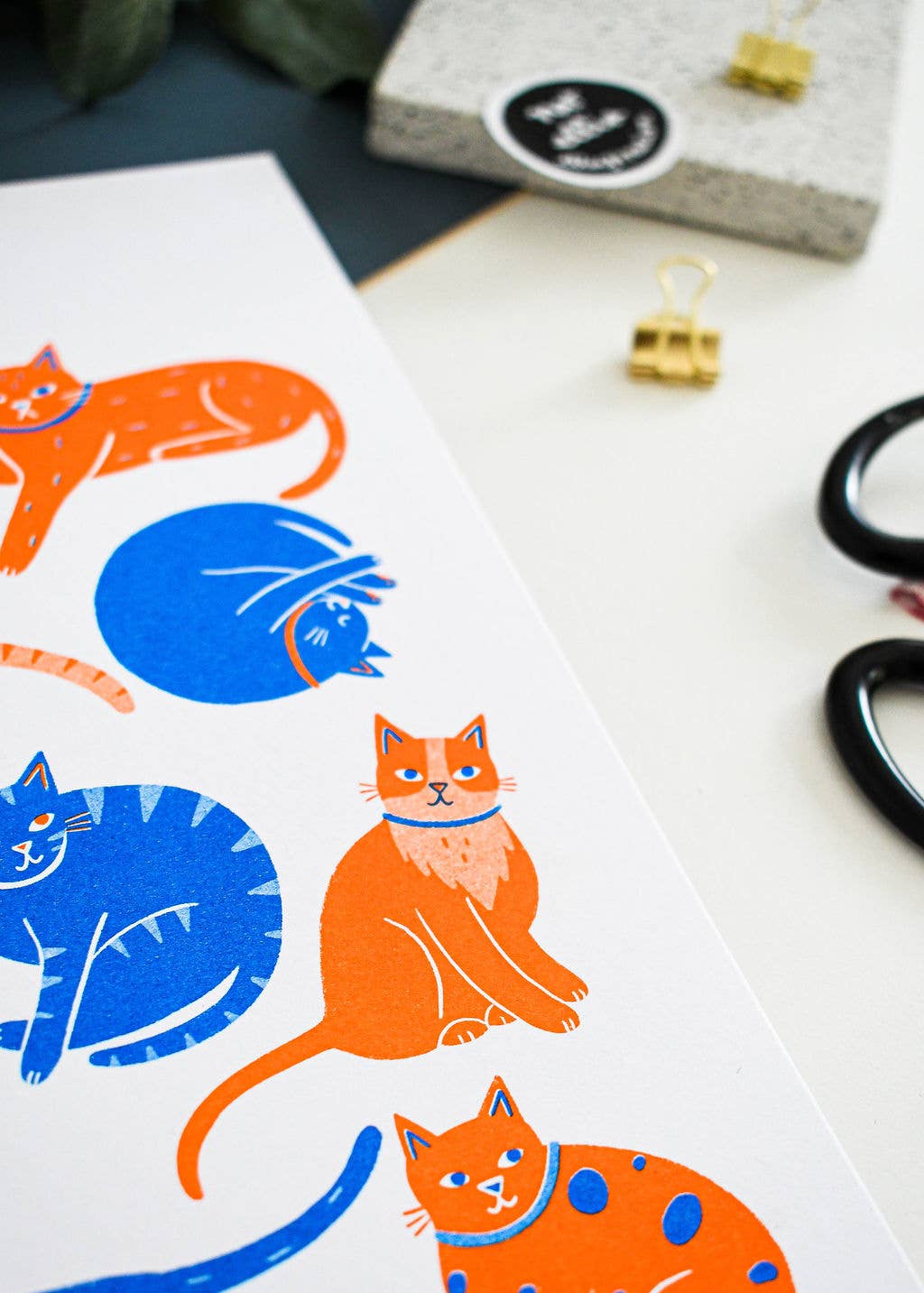 A close-up of a printed sheet featuring colorful cat illustrations in orange and blue, styled on a white desk with scissors, binder clips, and a small box in the background.