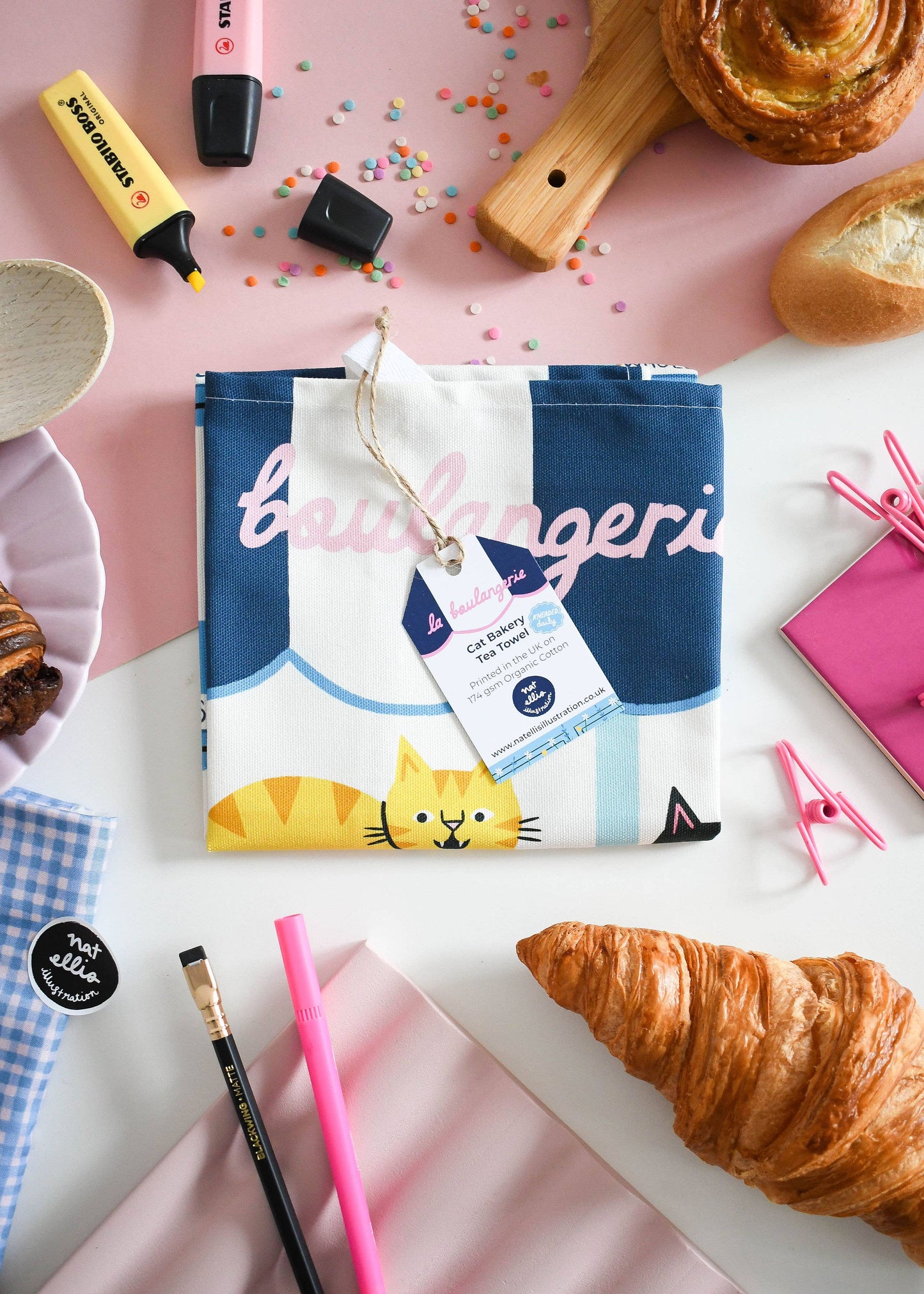 Product packaging with a cat design on a pink surface with pastries and stationery items.
