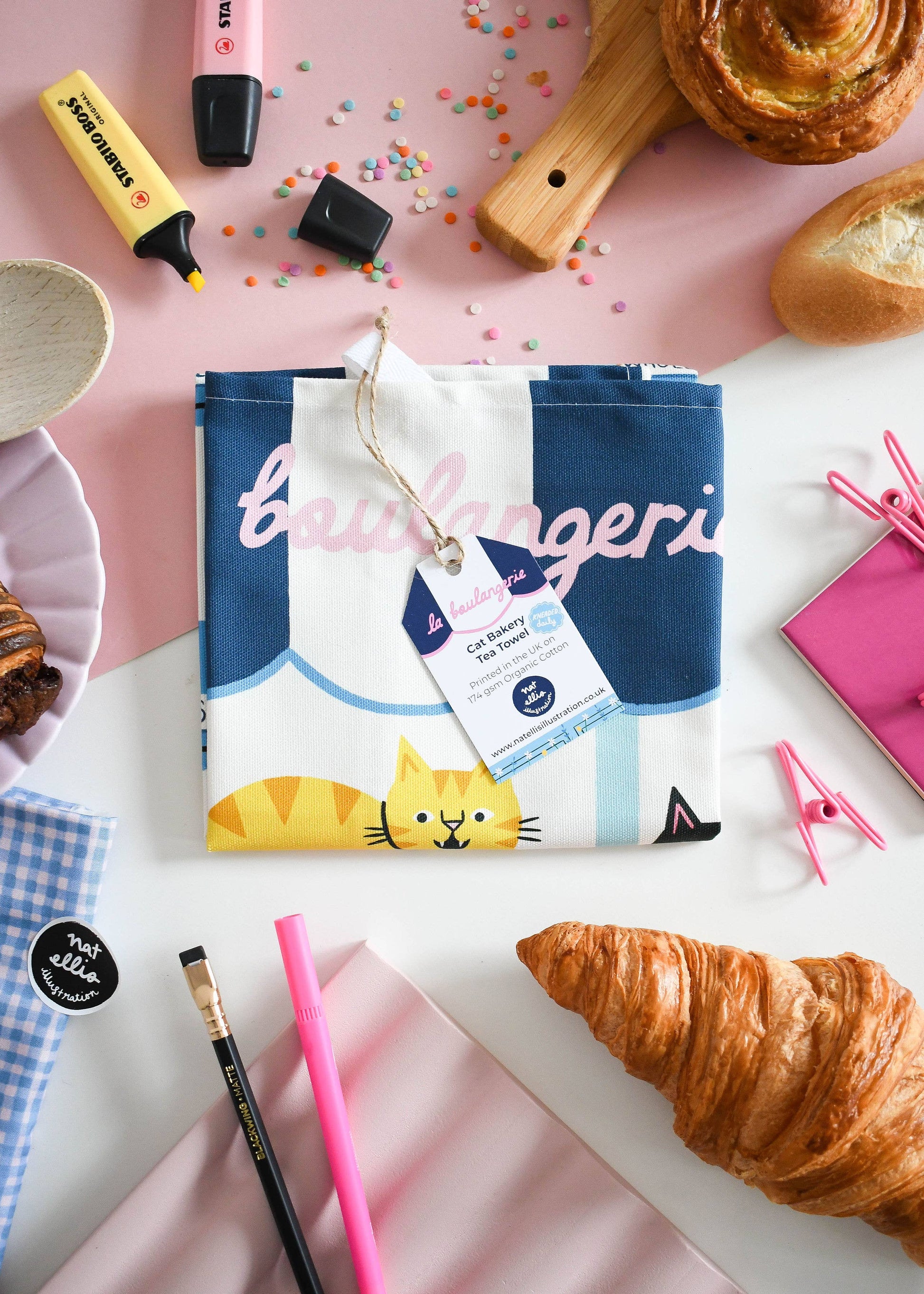 Product packaging with a cat design on a pink surface with pastries and stationery items.