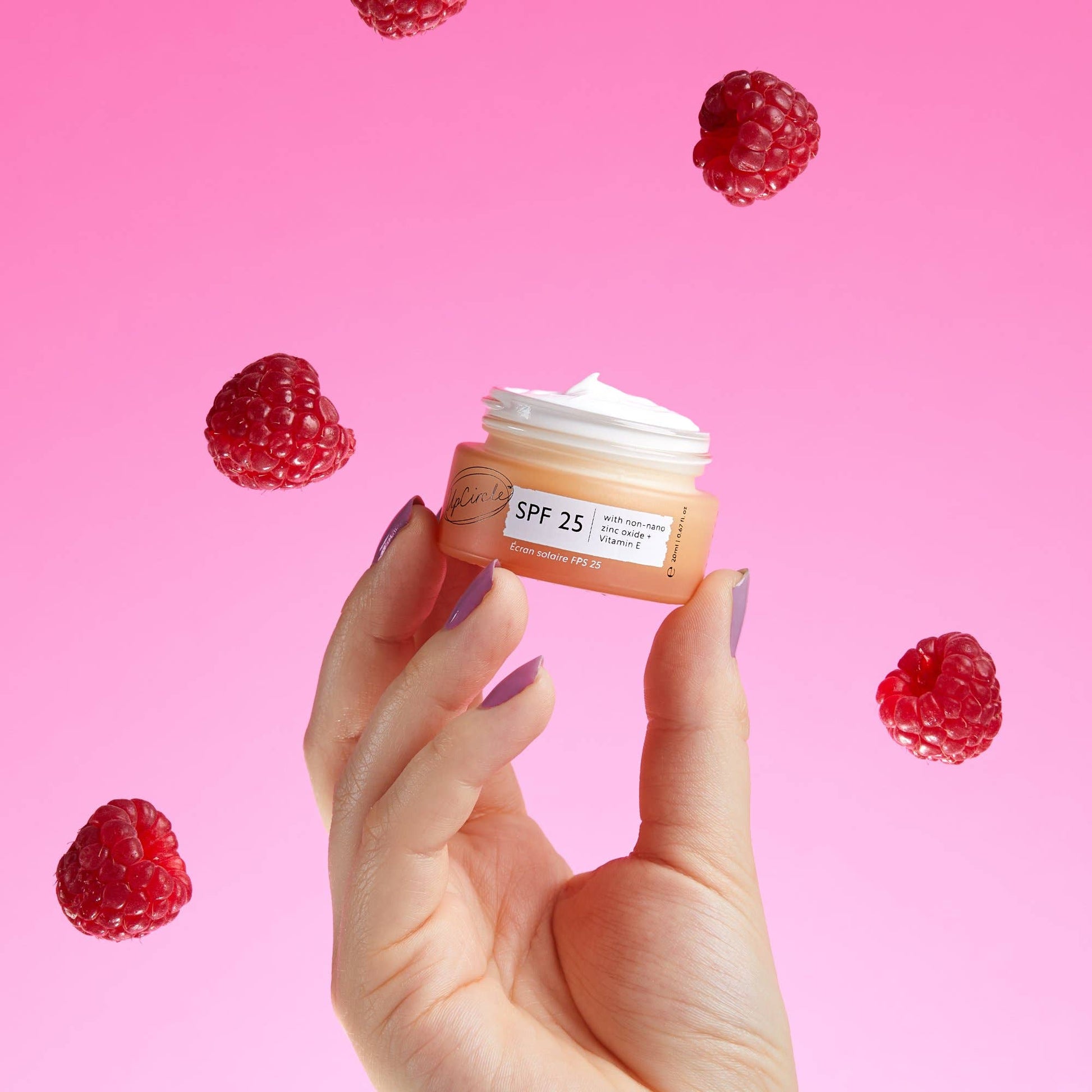 Hand holding a small jar of cream with raspberries on a pink background