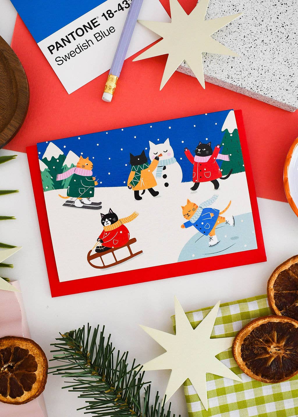 A square greeting card with a colorful winter scene featuring cartoon cats and a dog ice skating, surrounded by holiday-themed flat-lay props including dried orange slices, pine branches, paper stars, and a Pantone card on a red background.