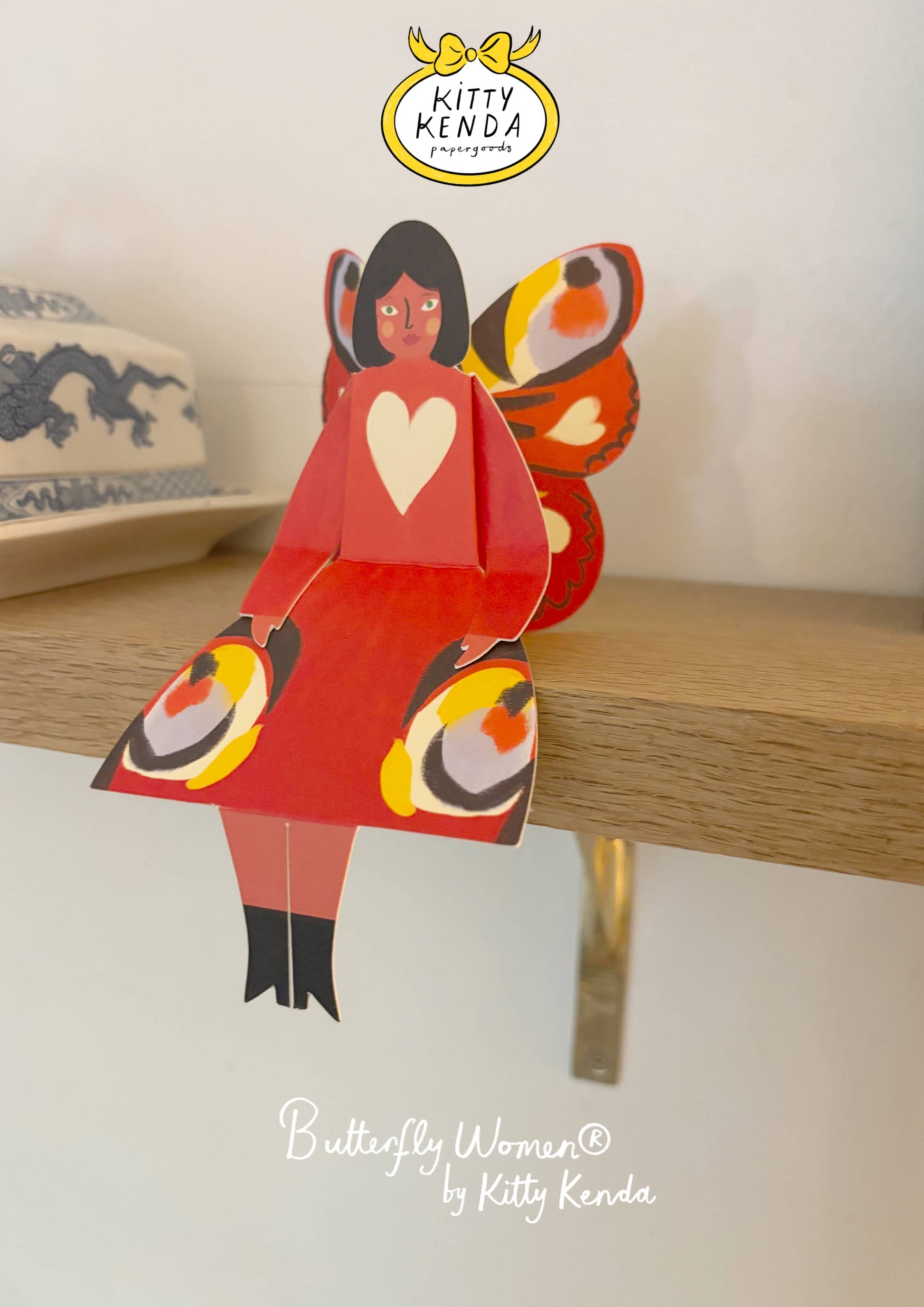 Colorful paper figure of a woman with butterfly wings on a wooden surface, branded 'Kitty Kenda'.