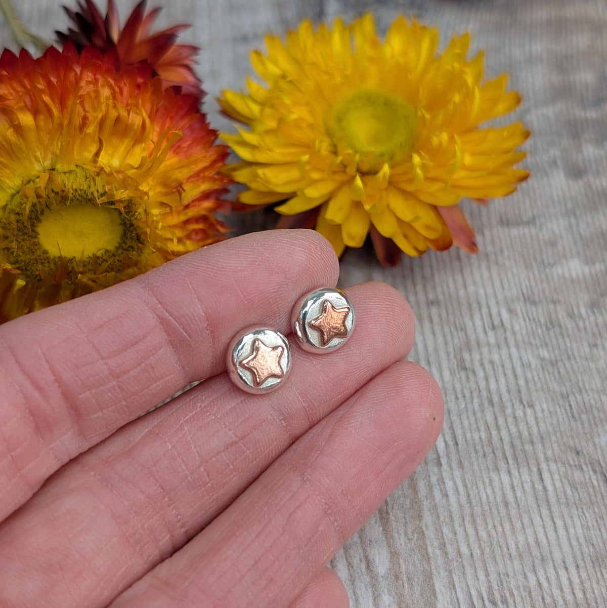 A pair of small round stud earrings with a silver-toned outer ring and a rose-gold-toned star-shaped center, shown resting on a person's fingers with yellow and orange flowers blurred in the background.