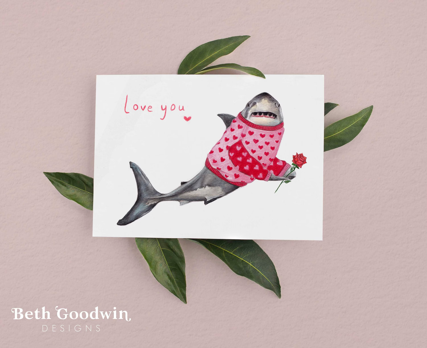 Card with a shark wearing a pink sweater holding a flower, on a pink background with green leaves.