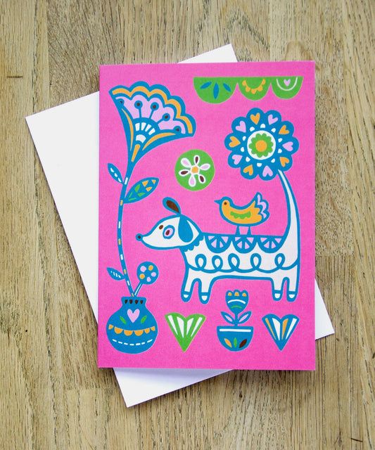 A colourful card with a dog and floral designs on a pink background, placed on a wooden surface.