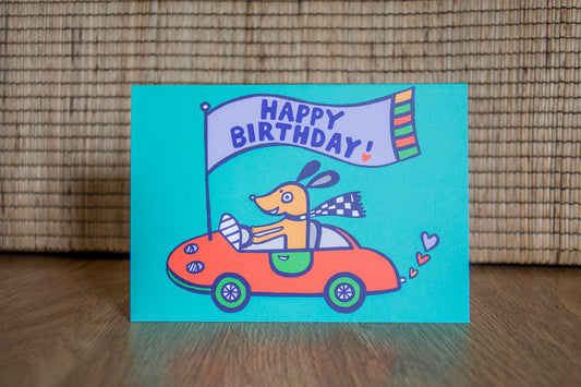 A card with a teal background depicting a happy brown dog in a red sports car, attached to the bonnet is a flag that reads "Happy Birthday!"