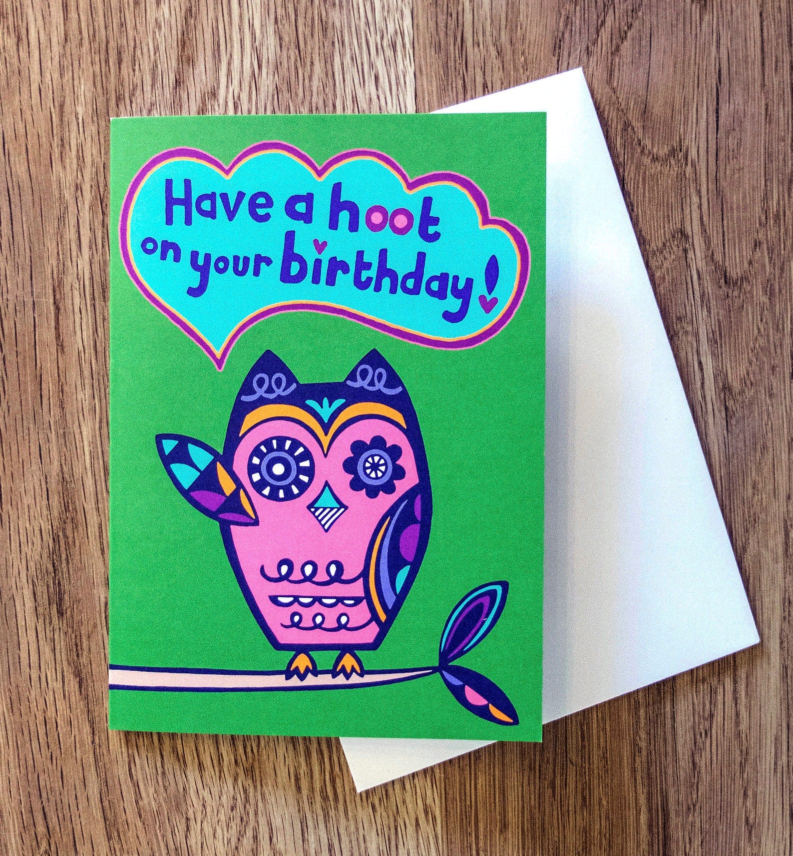 A green card with a pink and navy blue art deco style owl raising one of its wings, with a speech bubble that reads "Have a hoot on your birthday!"
