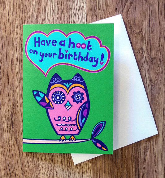 A green card with a pink and navy blue art deco style owl raising one of its wings, with a speech bubble that reads "Have a hoot on your birthday!"
