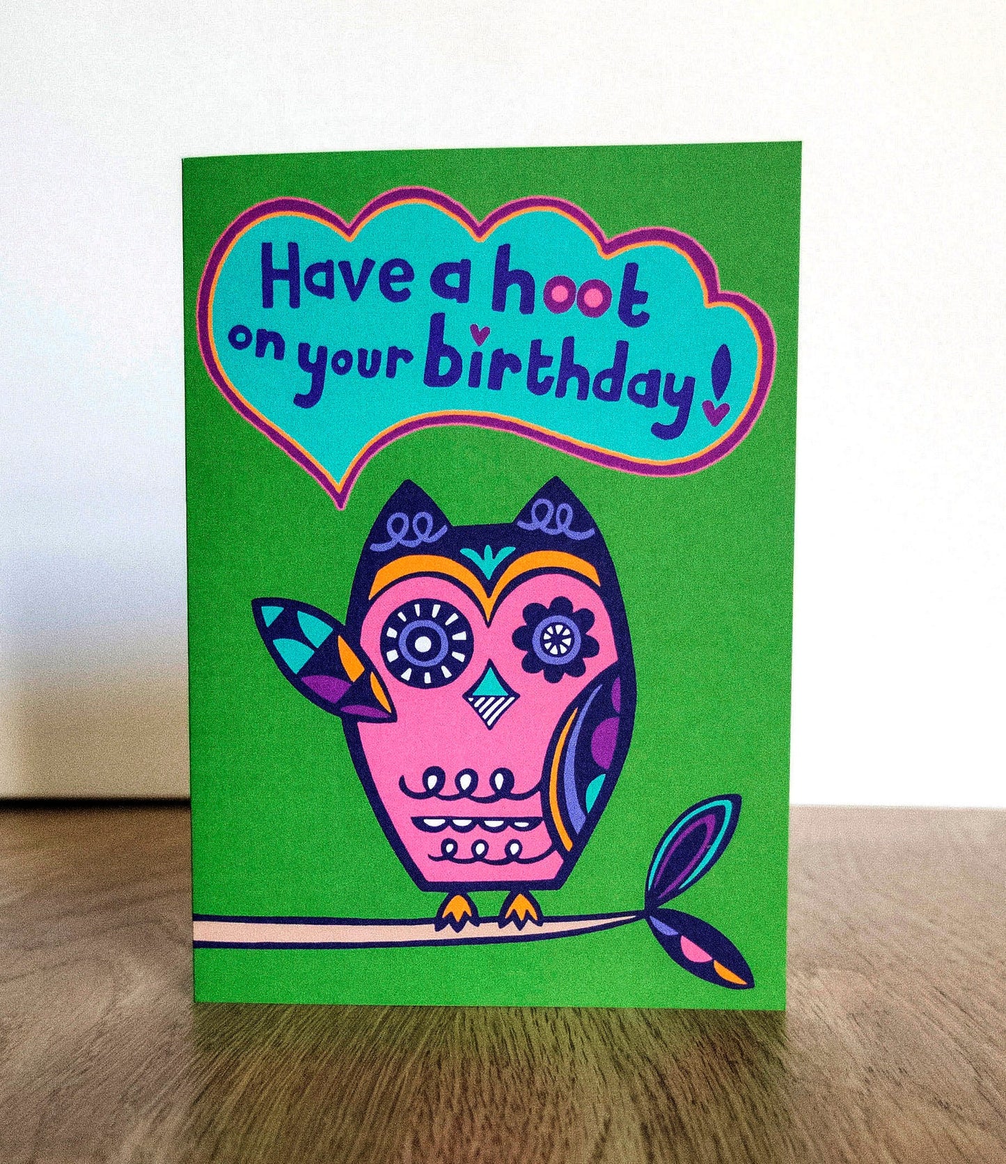 A green card with a pink and navy blue art deco style owl raising one of its wings, with a speech bubble that reads "Have a hoot on your birthday!"