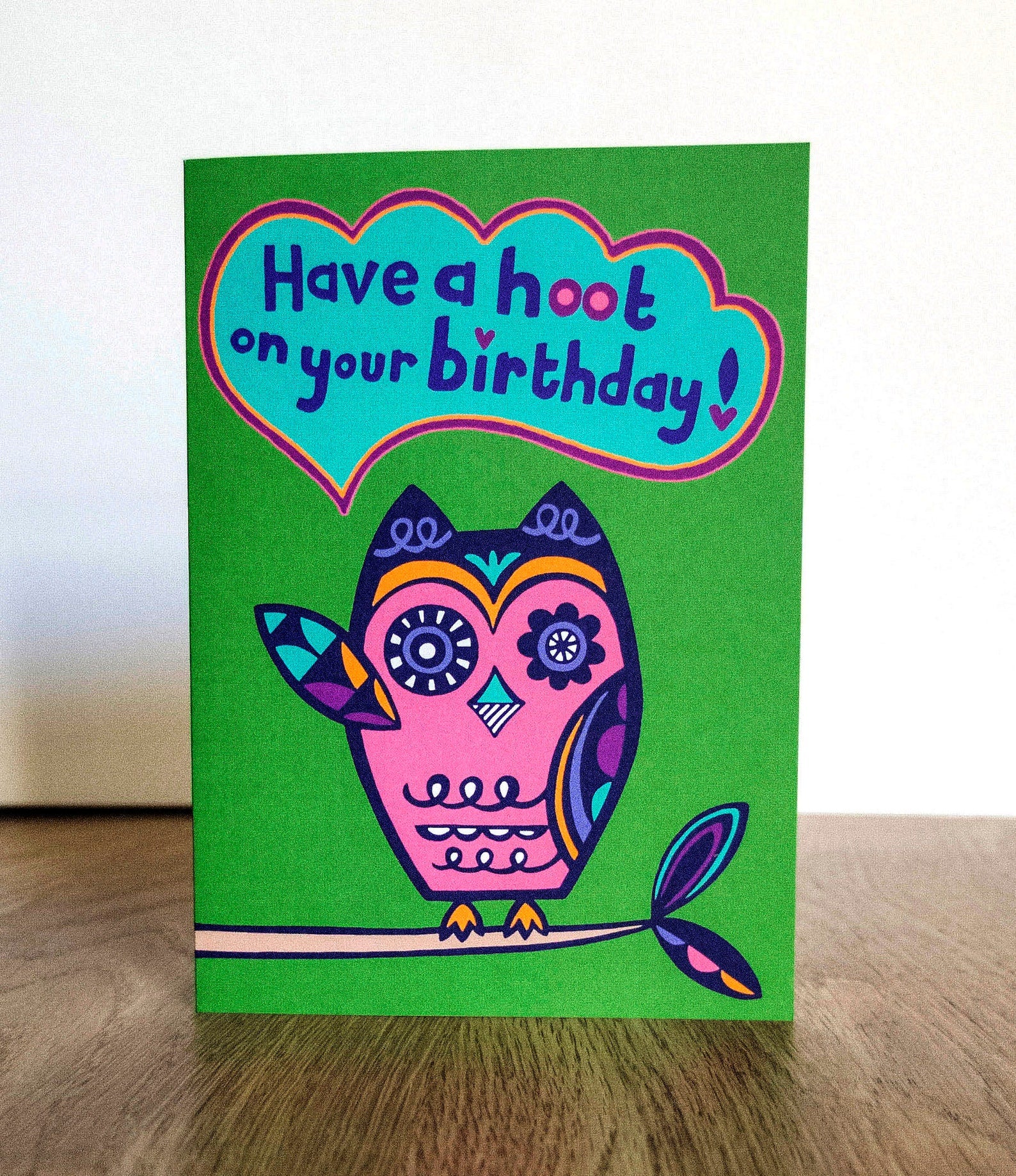 A green card with a pink and navy blue art deco style owl raising one of its wings, with a speech bubble that reads "Have a hoot on your birthday!"