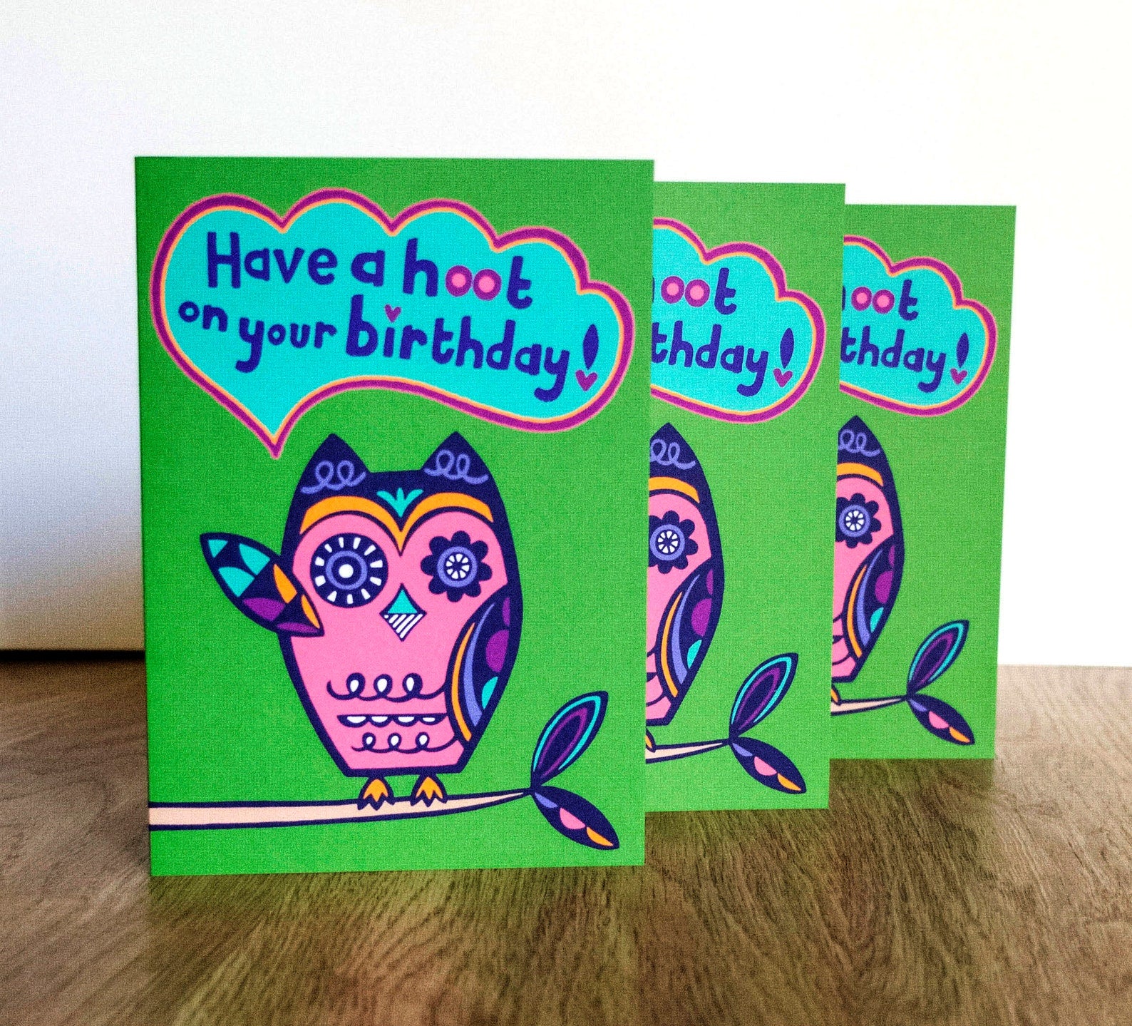 A green card with a pink and navy blue art deco style owl raising one of its wings, with a speech bubble that reads "Have a hoot on your birthday!"