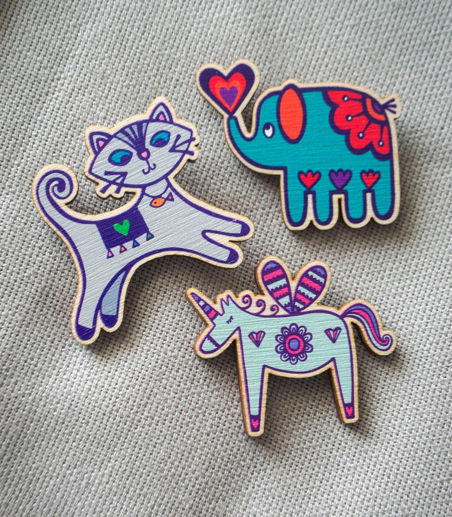 Three brightly painted wooden brooches.  Pictured are a grey leaping cat, a turquoise blue elephant and powder blue and pink unicorn.