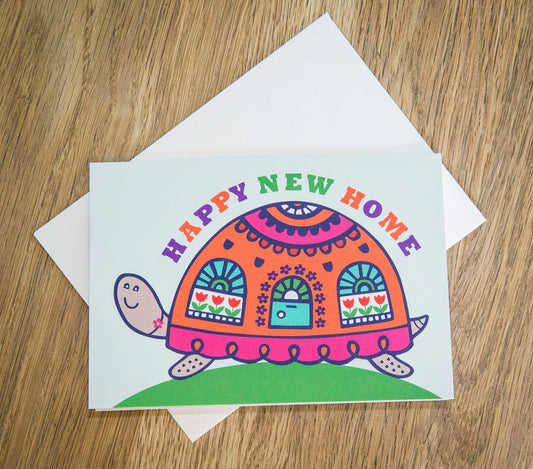 A card depicting a colourful happy tortoise with a door and windows on its brightly decorated shell. Curved above the shell reads the text "Happy New Home".