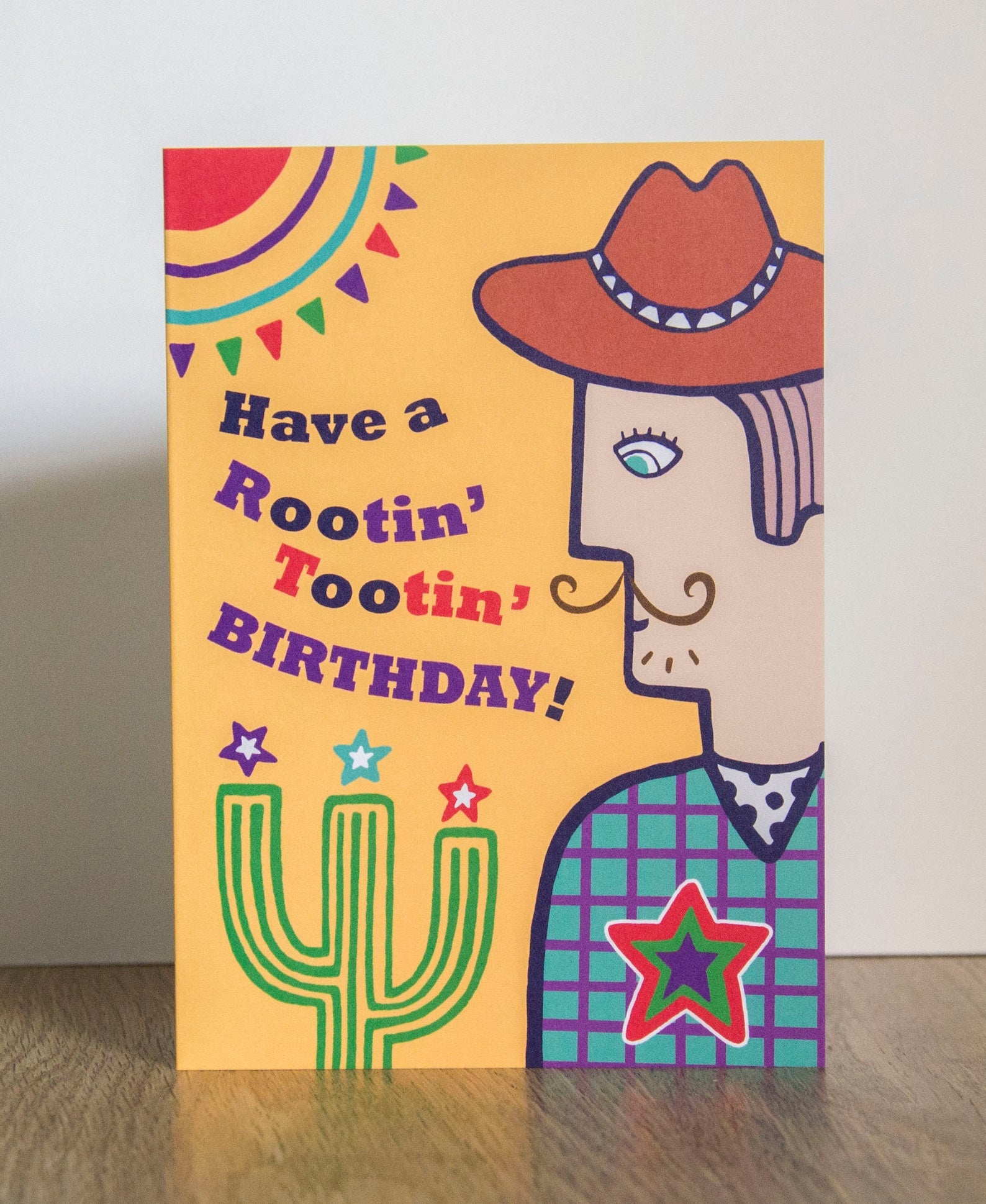 A card depicting a moustached cowboy next to a cactus, with a yellow background. To the left of the cowboy there is text that reads "Have a Rootin' Tooting' Birthday!"