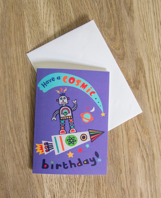 A vibrant card that depicts a robot standing atop a rocket in space. Above the robot there is a shooting star. The text on the card reads "Have a cosmic birthday!"