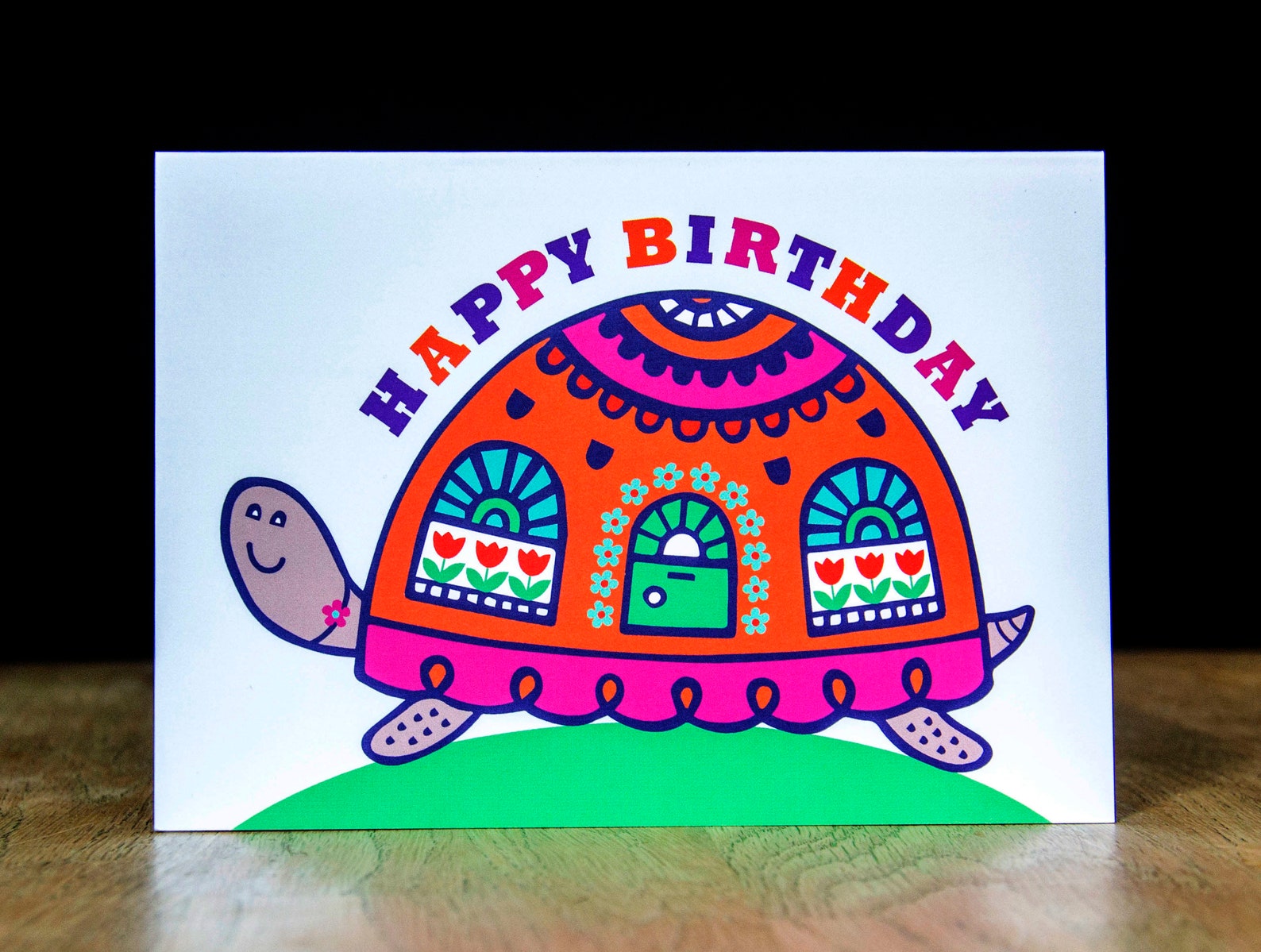 A card depicting a bright and colourful happy tortoise with a door and windows on its shell. Curved above the shell is the text "Happy Birthday".