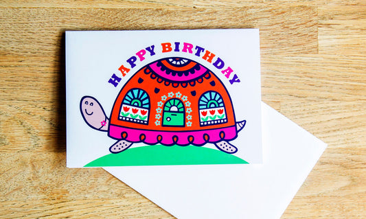 A card depicting a bright and colourful happy tortoise with a door and windows on its shell. Curved above the shell is the text "Happy Birthday".