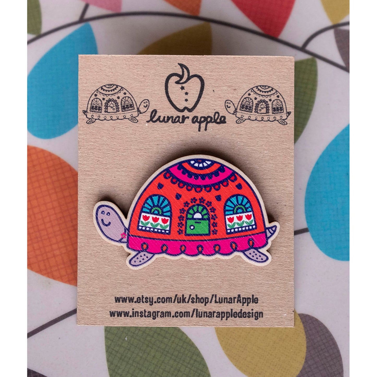 A brightly painted wooden brooch of an art deco inspired tortoise with windows and a door on its shell, pinned to a brown card backing.