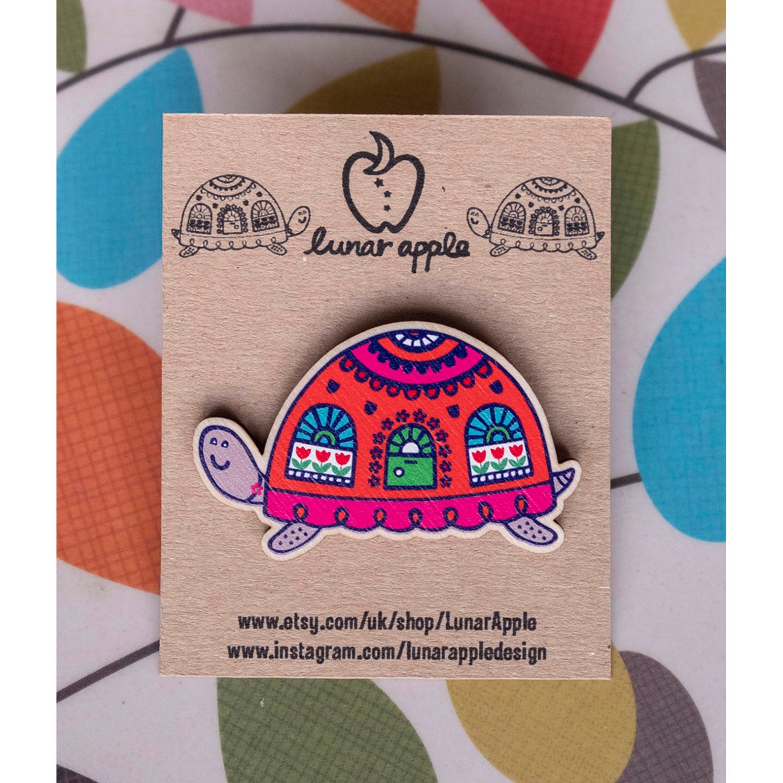 A brightly painted wooden brooch of an art deco inspired tortoise with windows and a door on its shell, pinned to a brown card backing.