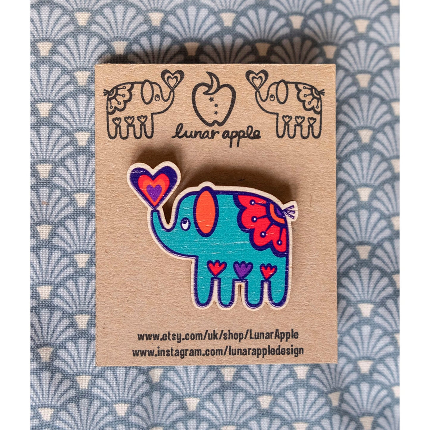 A brightly painted wooden brooch of an art deco inspired blue elephant pinned to a brown card backing.