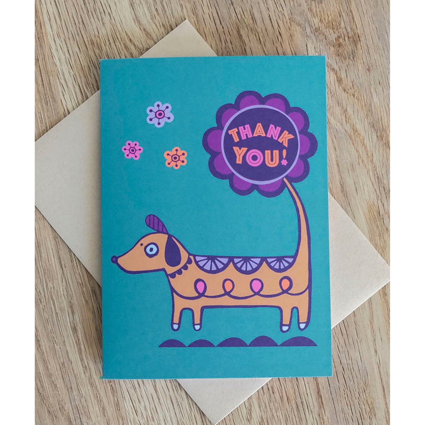 A teal thank you card with the phrase "Thank You!" in a purple flower at the end of a brown sausage dog's tail.