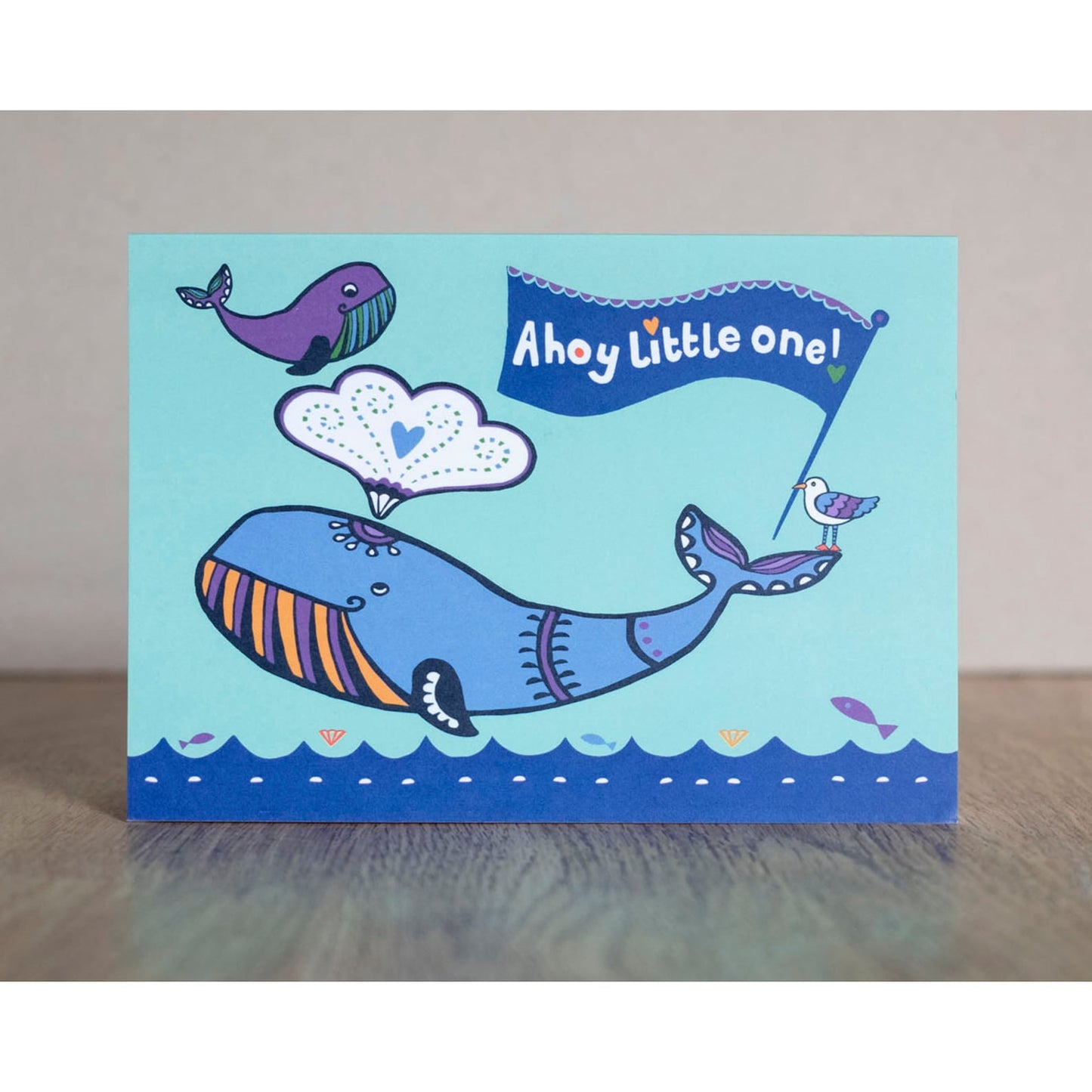 A blue card depicting an adult and baby whale above an ocean. On the adult's tail there is a seagull holding a flag in its beak. The flag reads "Ahoy little one!"