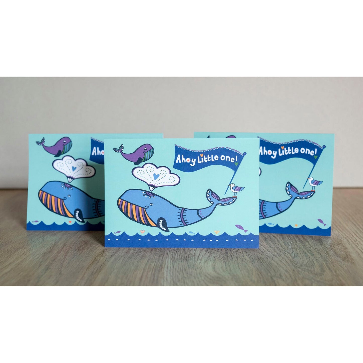 A blue card depicting an adult and baby whale above an ocean. On the adult's tail there is a seagull holding a flag in its beak. The flag reads "Ahoy little one!"