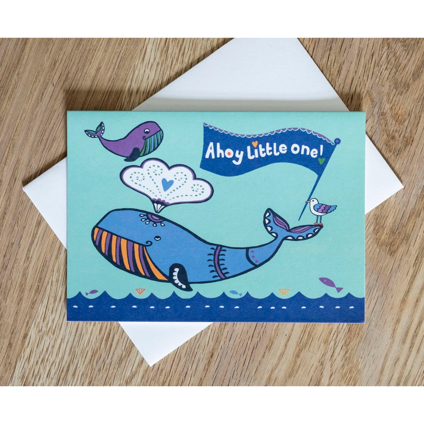 A blue card depicting an adult and baby whale above an ocean. On the adult's tail there is a seagull holding a flag in its beak. The flag reads "Ahoy little one!"