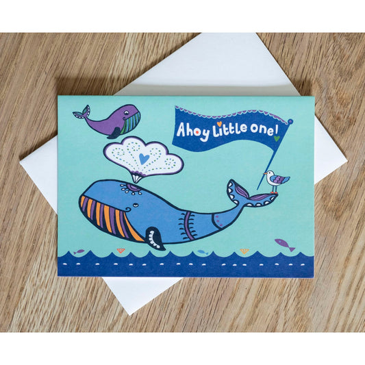A blue card depicting an adult and baby whale above an ocean. On the adult's tail there is a seagull holding a flag in its beak. The flag reads "Ahoy little one!"