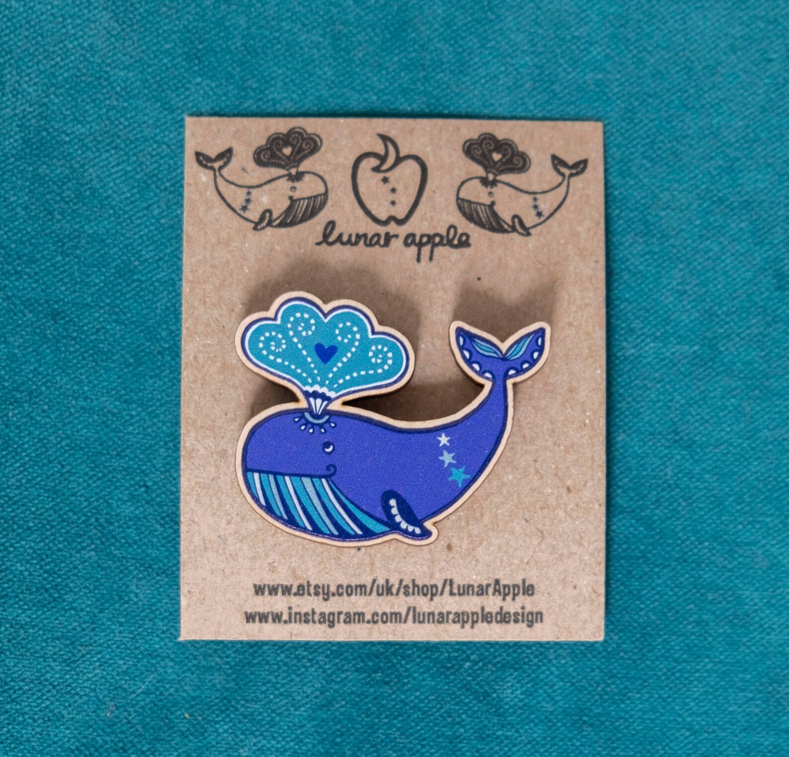 A wooden brooch of a cartoon whale pinned to a brown card backing.