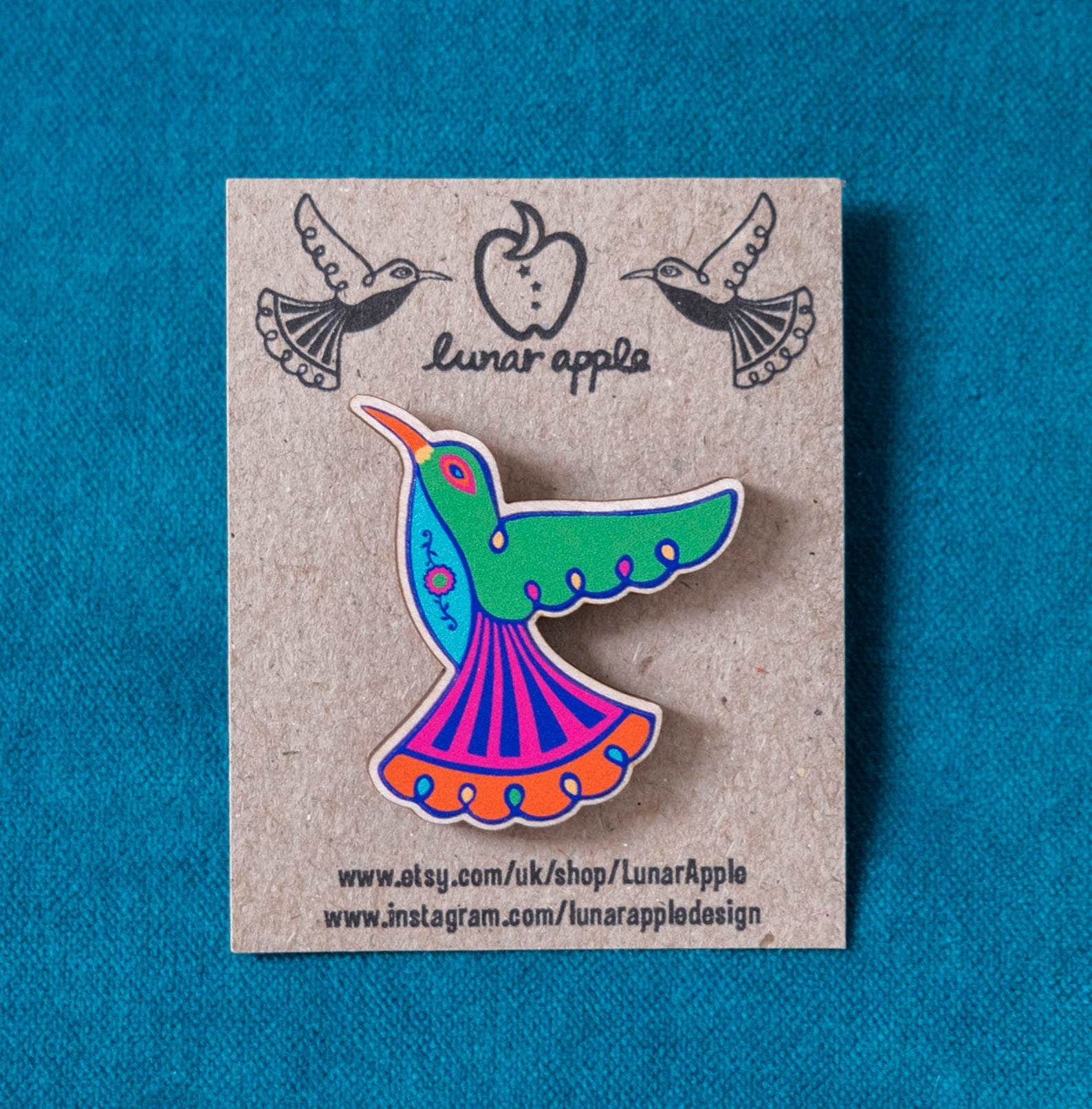 A brightly painted wooden brooch of an art deco inspired hummingbird pinned to a brown card backing.