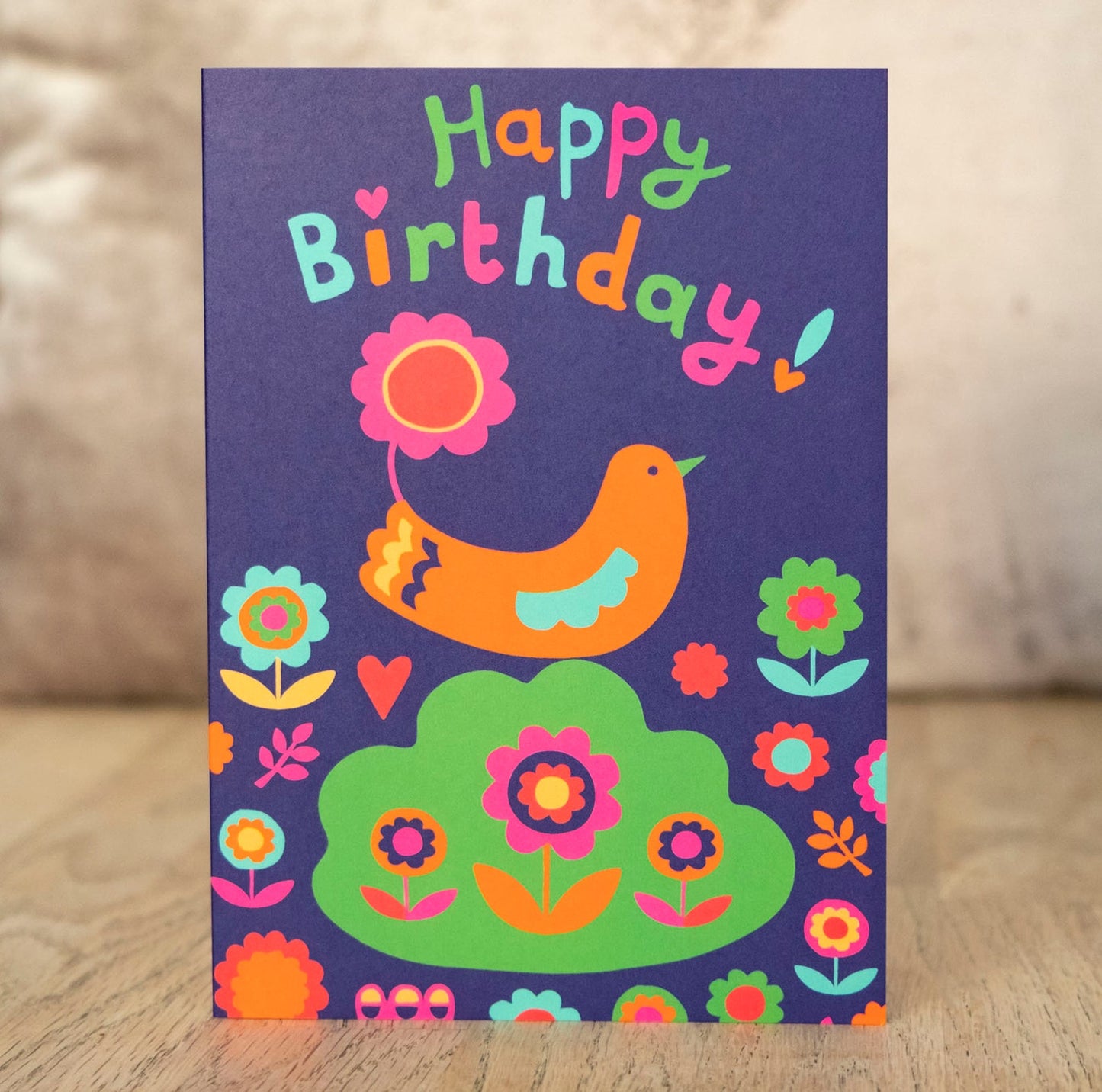 A dark blue card with the phrase "Happy Birthday!" in multicolour text. Underneath is a bird with a flower on its tail sitting on top of a shrub, surrounded by flowers.