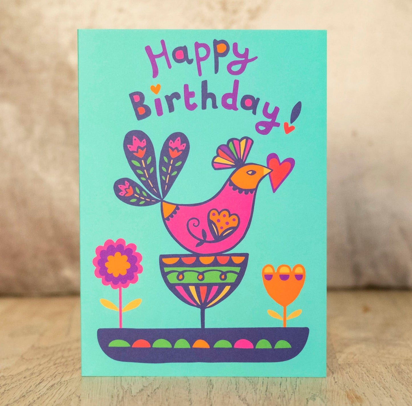 A turquoise card that says "Happy Birthday!" at the top. Underneath is a bird with a heart held in its beak, and flowers.