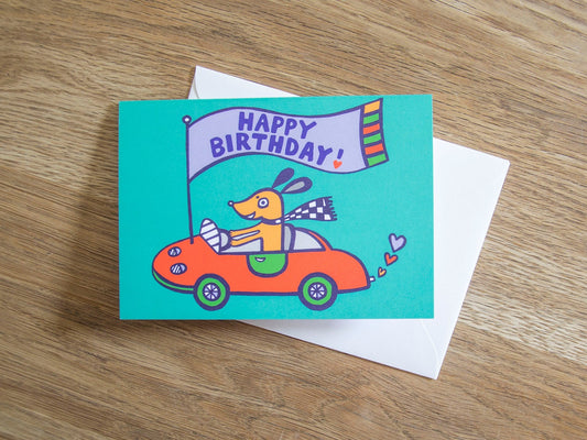 A card with a teal background depicting a happy brown dog in a red sports car, attached to the bonnet is a flag that reads "Happy Birthday!"