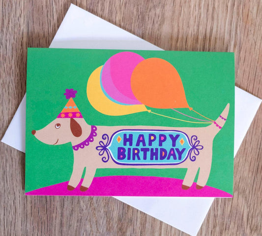 A card depicting a smiling sausage dog with balloons attached to its tail, with the message "happy birthday" across the body.