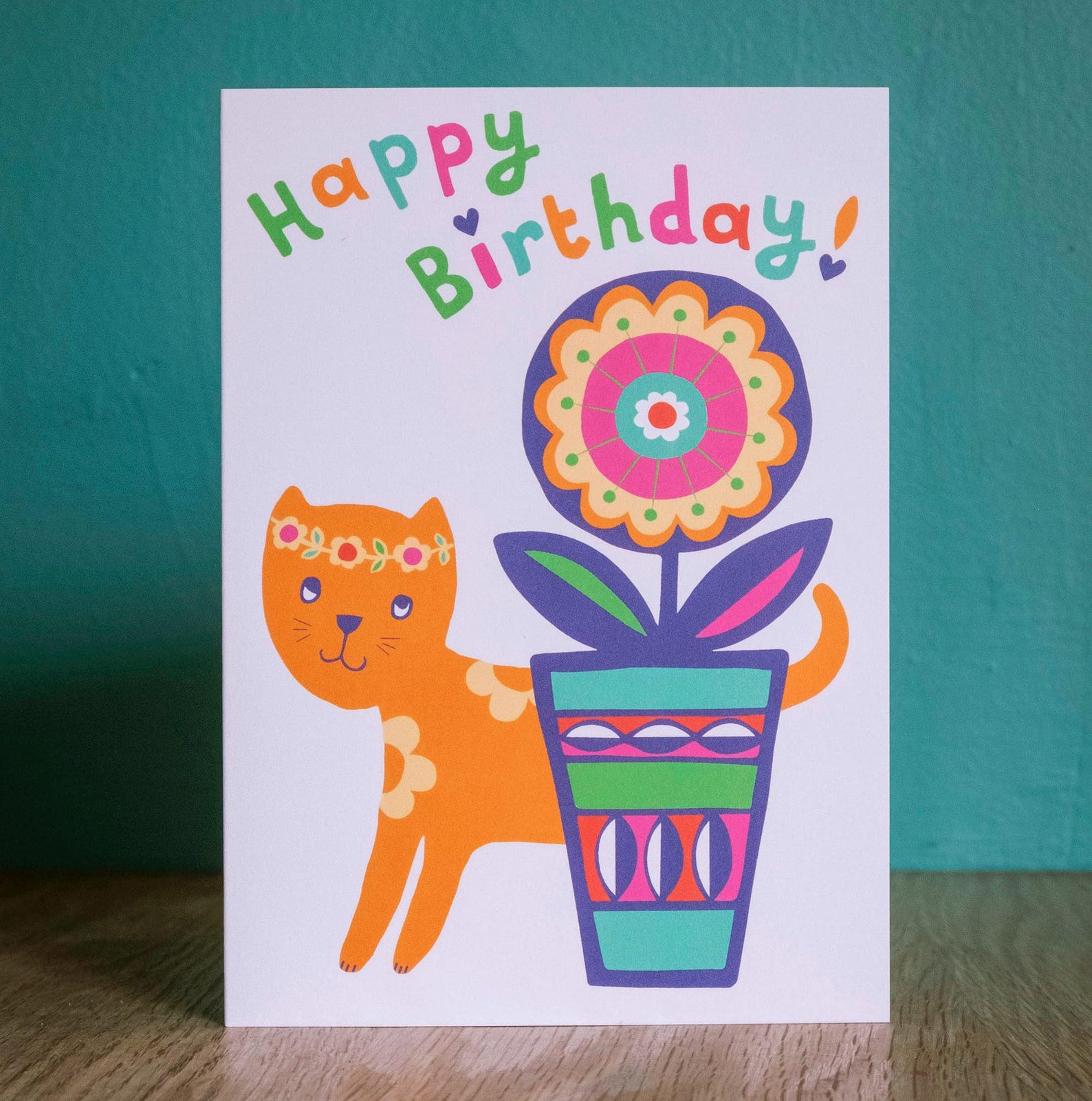 A pale pink card with the text "Happy Birthday!" in multicolour writing. Underneath is a ginger cat wearing a flower crown hiding behind a large flower in a flower pot.