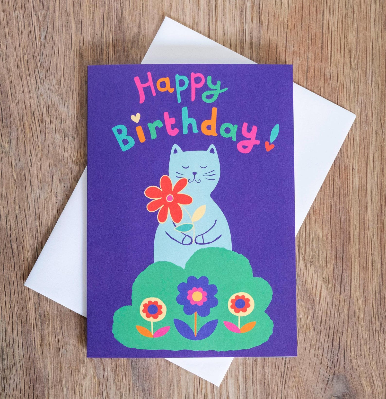 A blue card with the text "Happy Birthday!" in multicoloured writing. Underneath is a grey cat in a shrub holding a red flower.