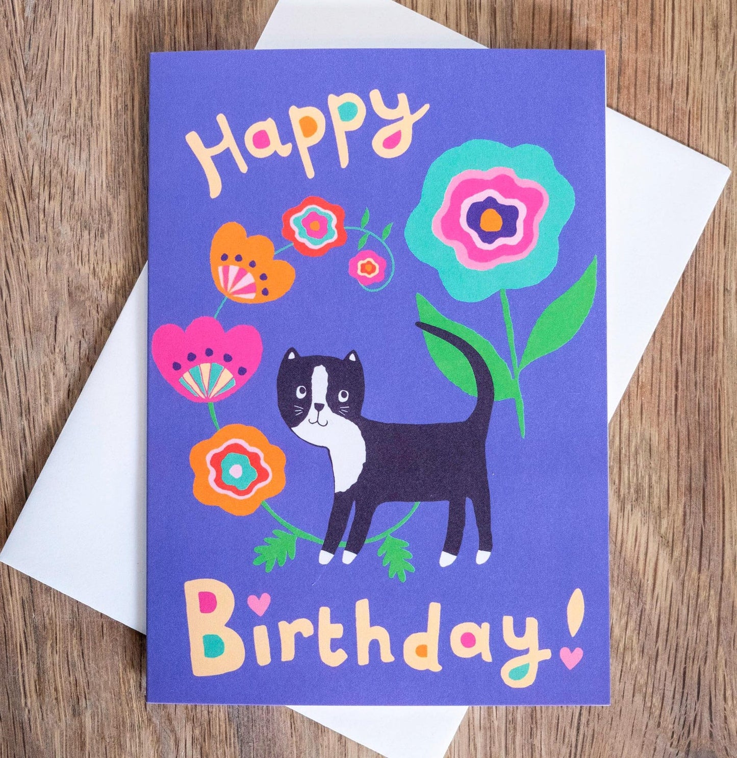 A blue card with the text "Happy Birthday!" on the top and bottom of the card. In the middle of the text there is a design of a black and white cat next to a swirling flower vine and large blue flower.