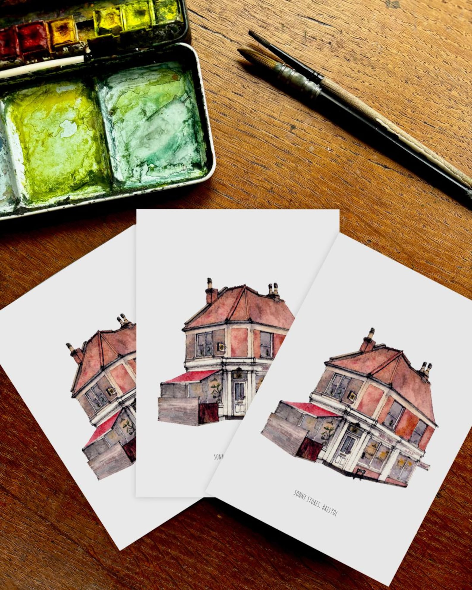 Watercolor painting of a house on a card with watercolor set and brush on wooden surface