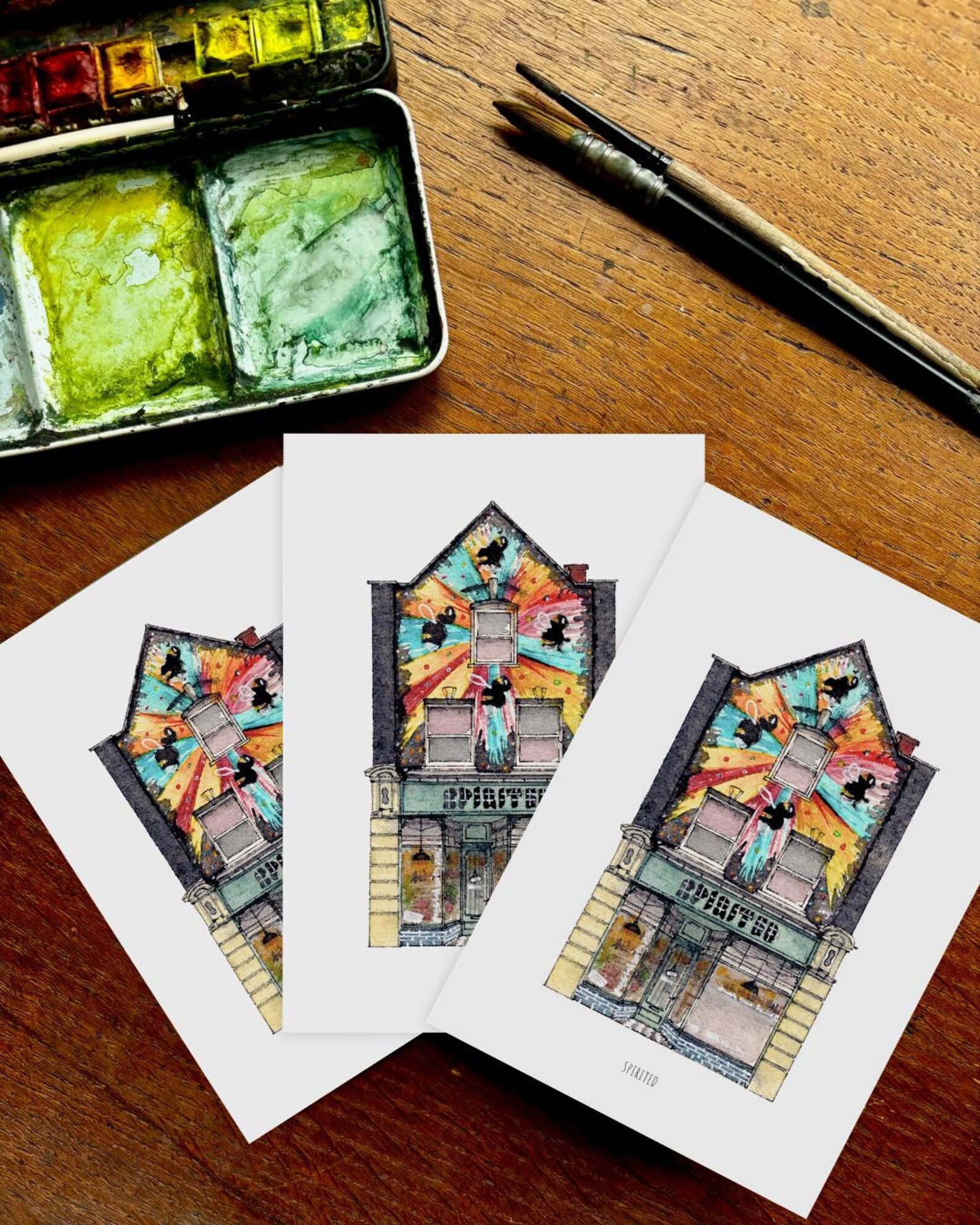 Colorful illustrations of buildings on white paper with watercolor set and brush on wooden surface