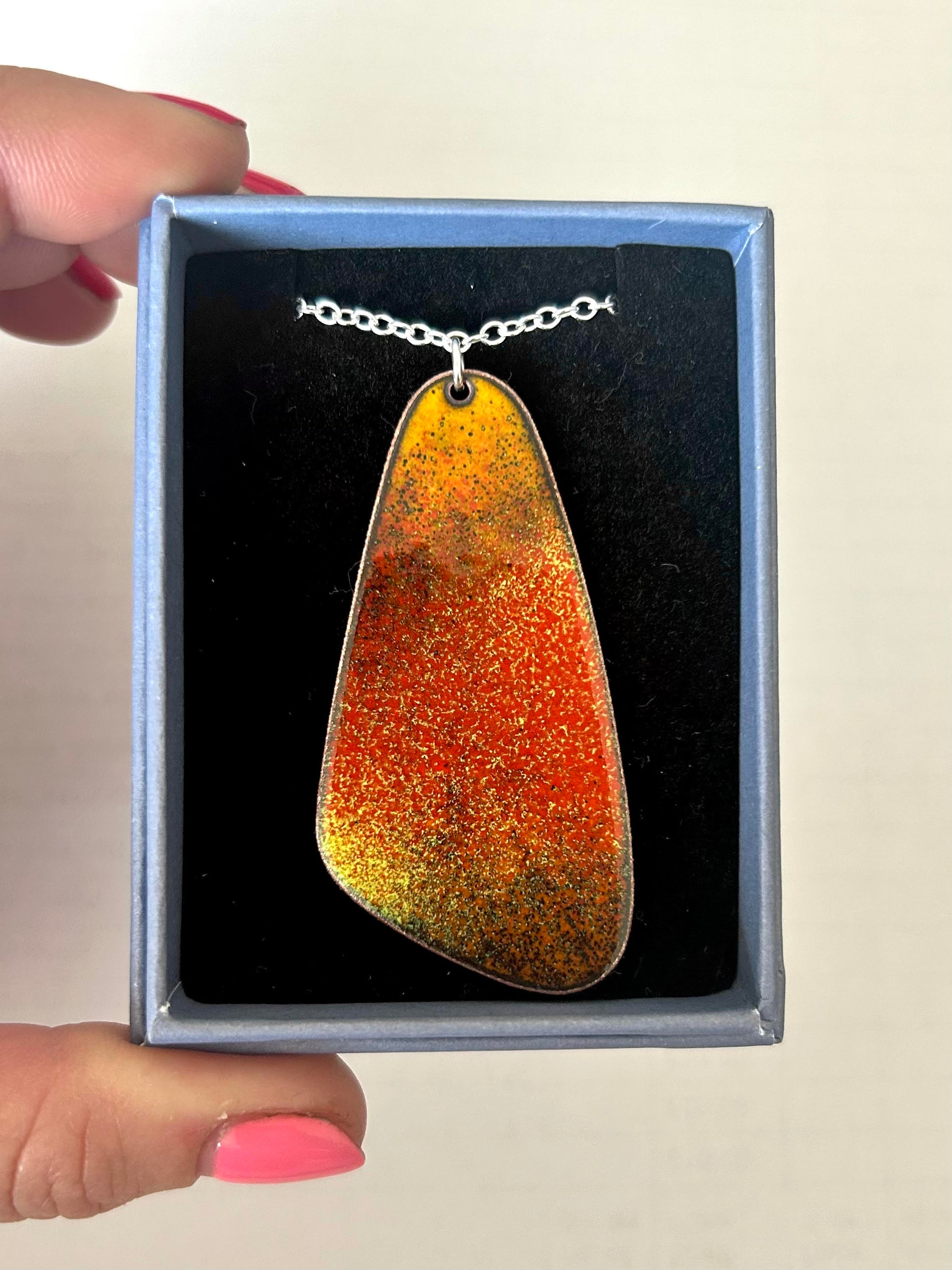 A pebble shaped orange and yellow ombre necklace with a silver chain, resting in a jewellery box.