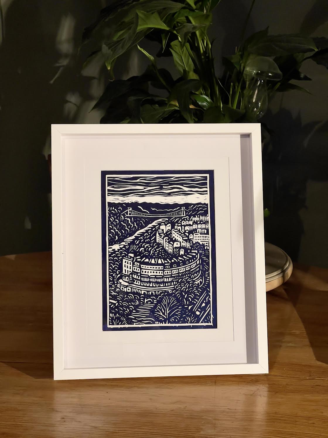 Framed artwork of a blue and white landscape on a wooden surface with a plant in the background.