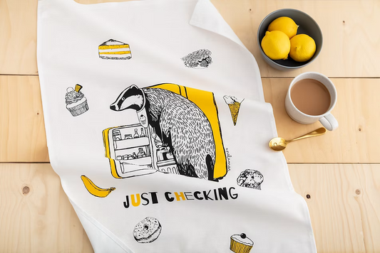 Tea towel with badger illustration and text 'Just Checking' on a wooden table with a cup of coffee and lemons.