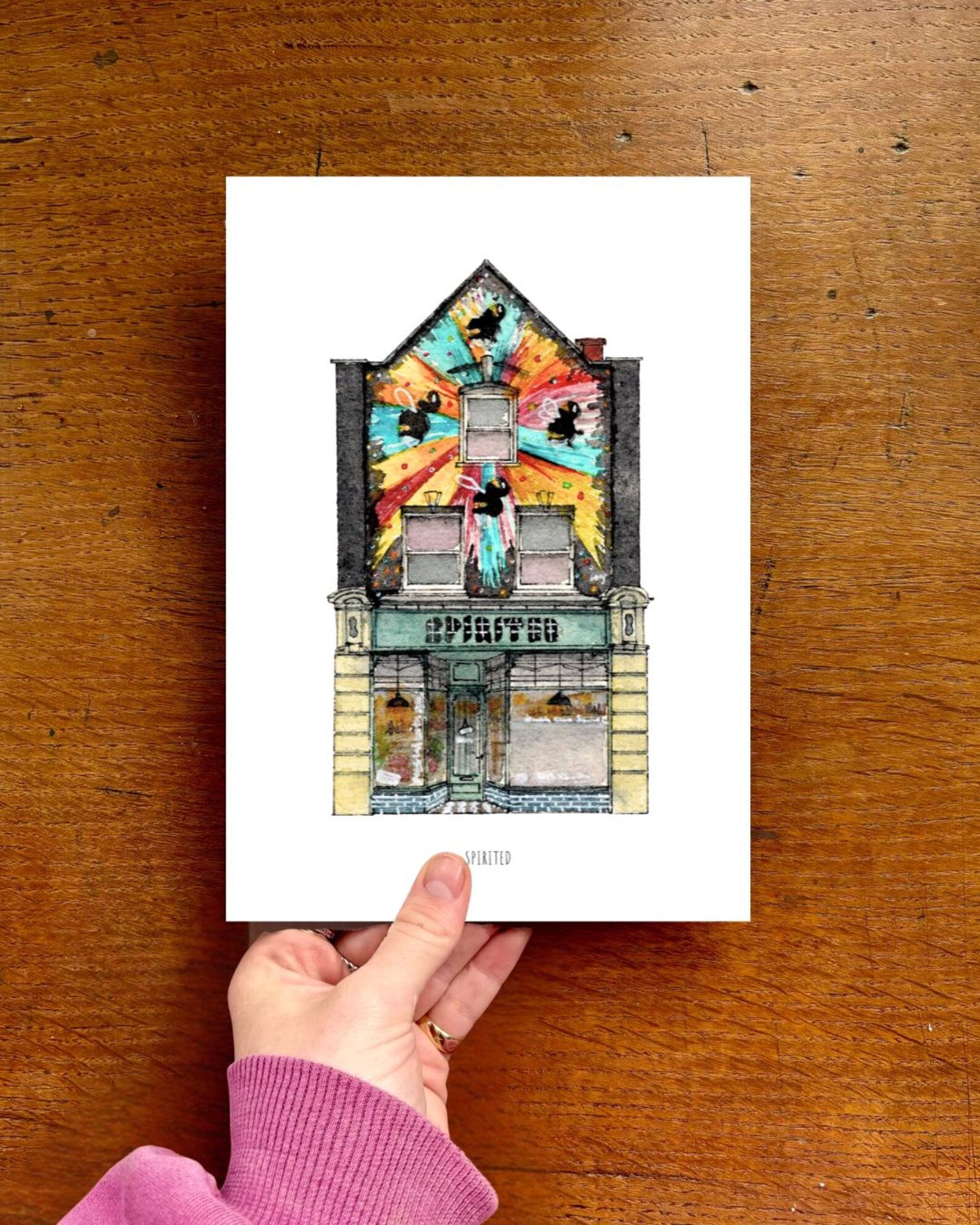 Hand holding a card with a colorful illustration of a building on a wooden surface